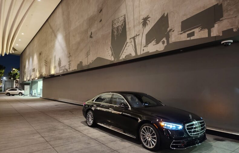 A sleek black Mercedes-Benz S-Class is parked under a modern overhang, with a large, shadowy mural in the background depicting urban scenes.