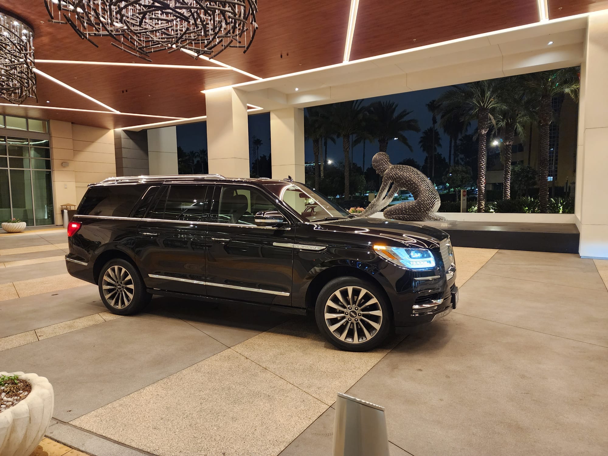 A sleek black SUV parked at a modern hotel entrance, featuring a striking wire sculpture and palm trees in the background under evening lights.