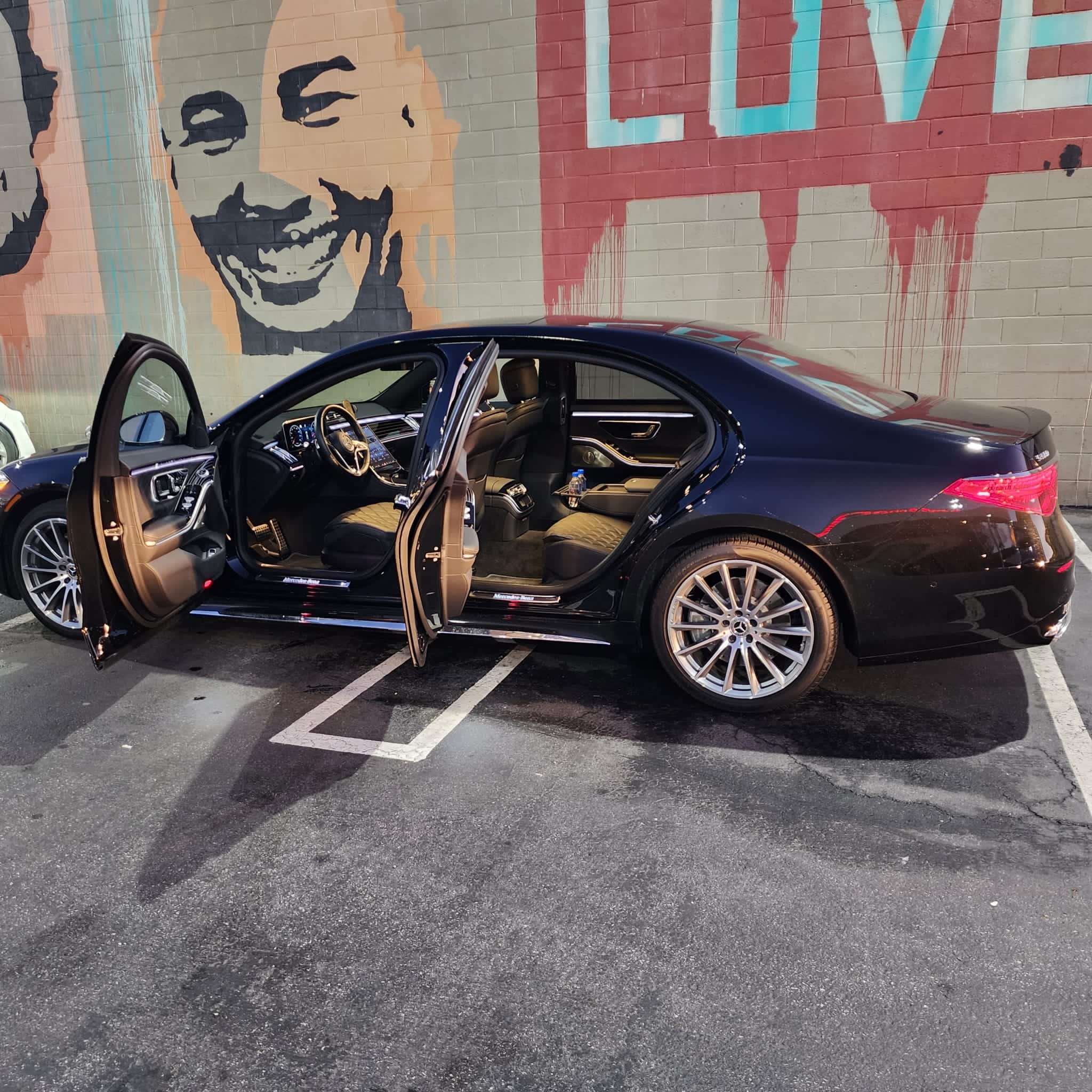 A black luxury sedan parked with both front doors open, showcasing its spacious interior, against a colorful mural featuring smiling faces.