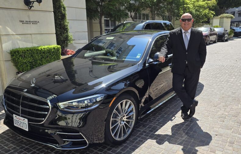 A man in a suit stands beside a black Mercedes-Benz S-Class outside the Peninsula Beverly Hills. The image showcases luxury and sophistication.
